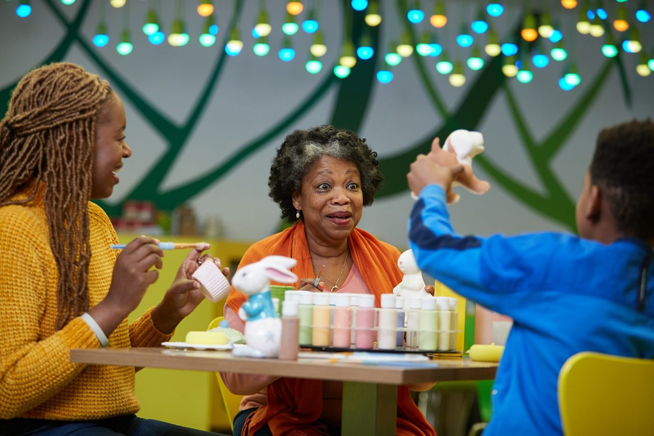 Three people paint ceramic bunnies at a table, chatting and smiling. Small paint jars and brushes arranged before them. Colorful string lights glow overhead in a bright craft studio.